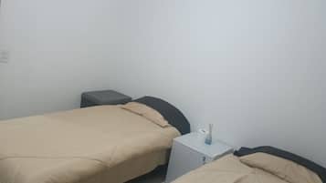 Ein kleines, schlichtes Zimmer mit zwei Betten, einem kleinen Tisch und einer weißen Wand.