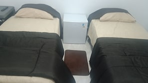 Una habitación con dos camas, una mesita y un electrodoméstico blanco en el centro.