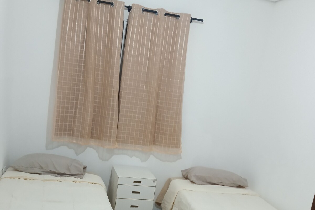 Una habitación pequeña y limpia con dos camas individuales, una mesita de noche y un armario pequeño.