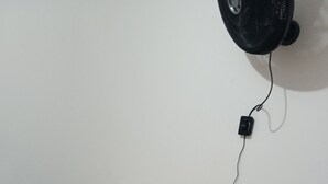 Uma cama de solteiro com lençol branco em quarto com parede branca e ventoinha preta fixada na parede.