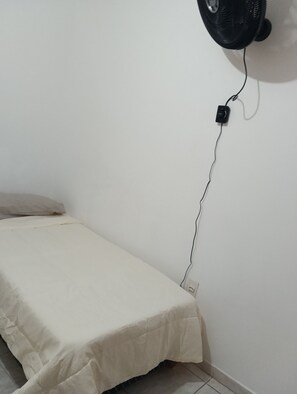 Una cama individual con sábana blanca en una habitación con pared blanca y un ventilador negro fijado a la pared.