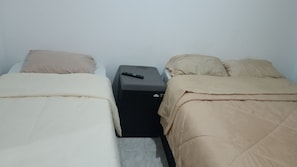Una habitación pequeña y sencilla con dos camas, cada una con una manta y almohada color beige, y una mesita de noche negra entre ellas.