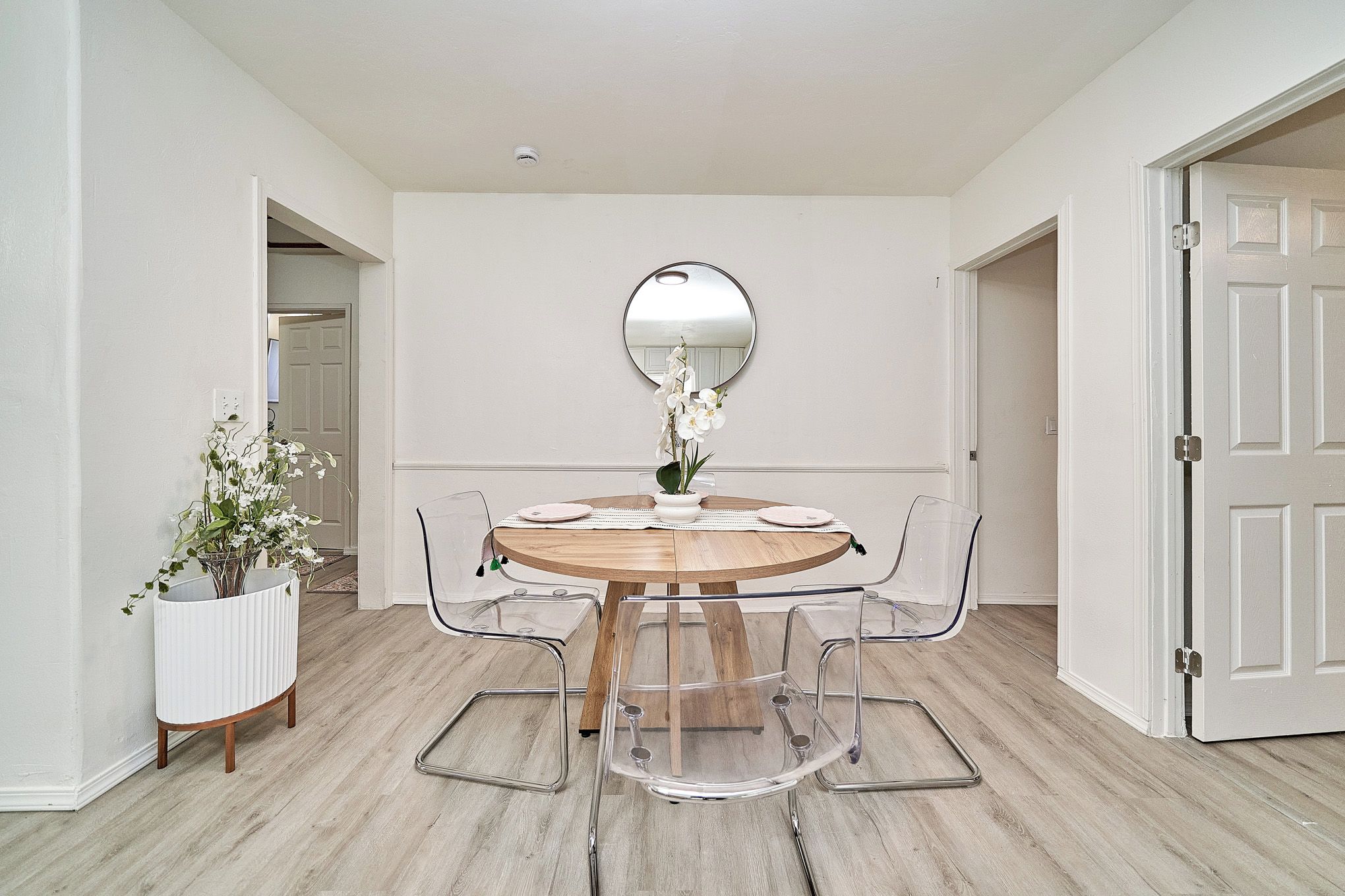 Un comedor con una mesa redonda, sillas transparentes, una planta en maceta y un espejo en la pared.