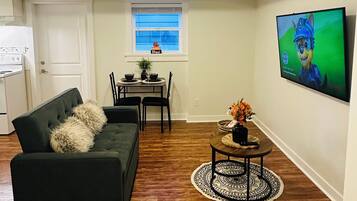 Een woonkamer met een donkergroene bank, een klein rond tafeltje met een vaas, een eethoek met een tafel en stoelen, en een keuken met een kookplaat en oven.