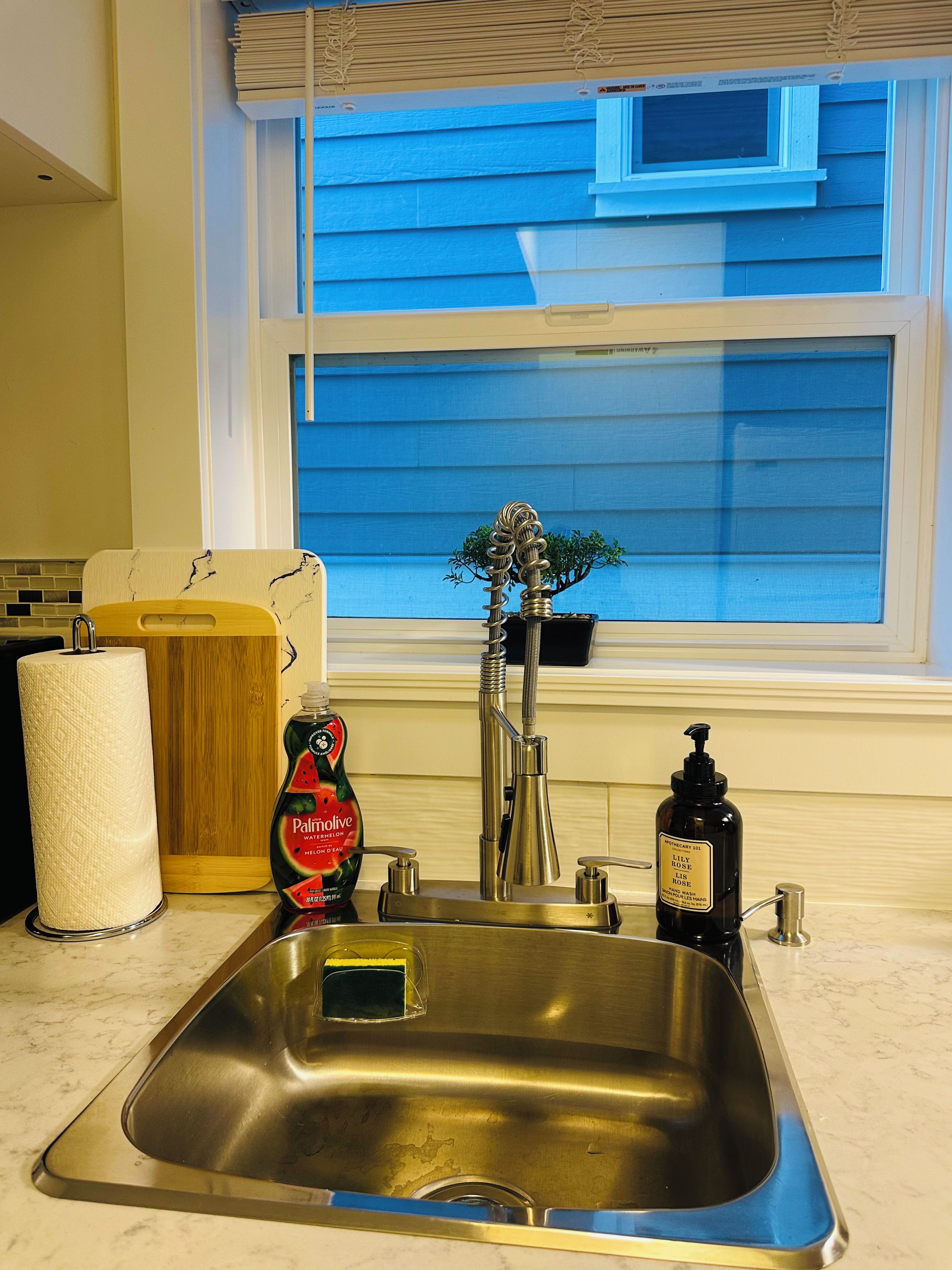 Een keuken met een roestvrijstalen spoelbak, een houten snijplank, een keukenrolhouder, een fles afwasmiddel en een raam met jaloezieën.