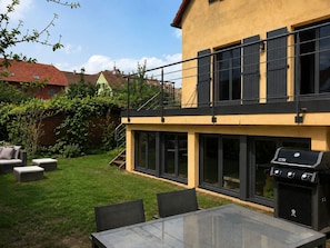 A modern two-story house with a balcony, a glass table, and a barbecue grill. - Single-family home with a garden—Alsace Wine Route—near Colmar. (Obermorschwihr)