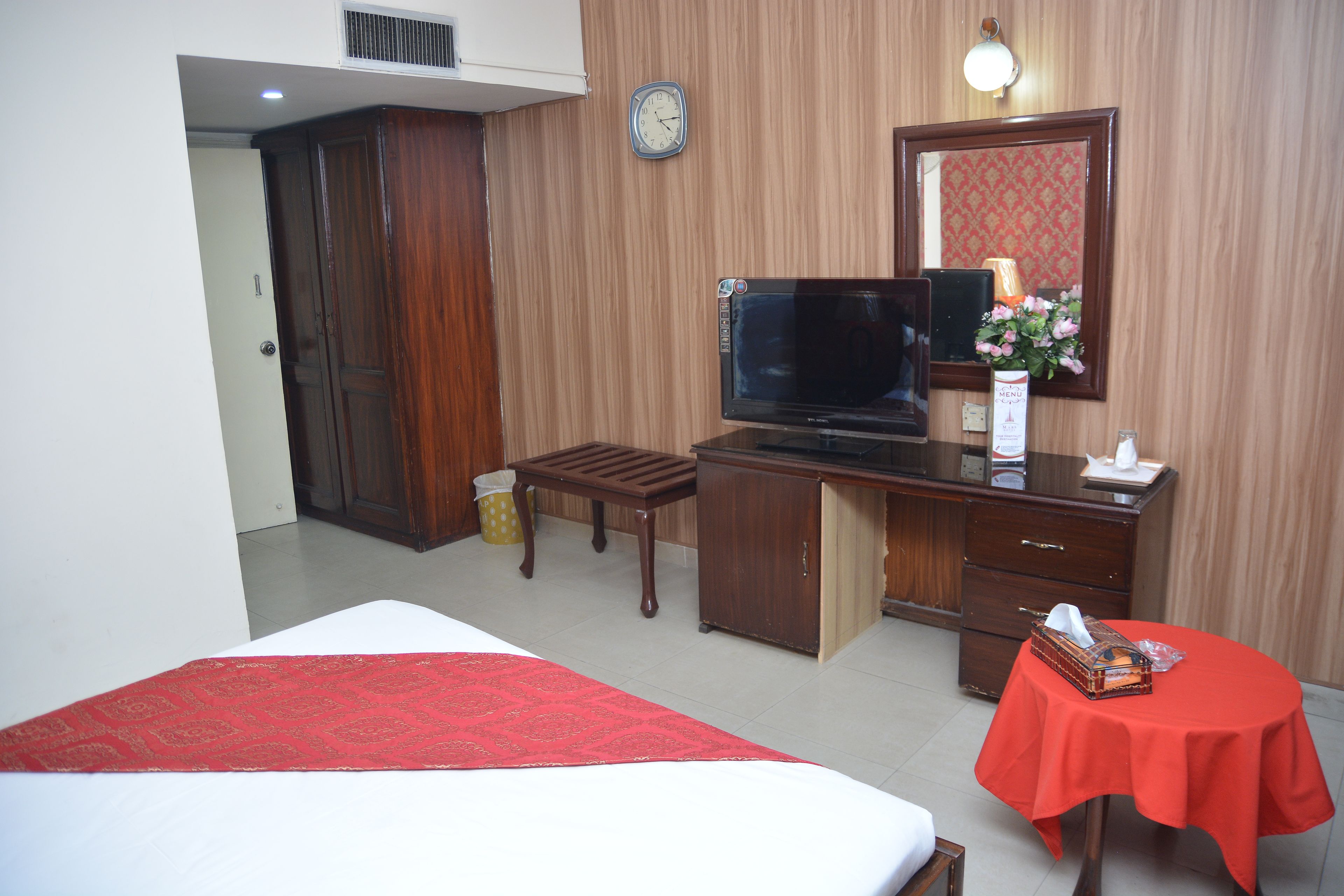Een hotelkamer met een bed, een bureau met een televisie, een spiegel, een klein tafeltje met een rode tafelkleed en een prullenbak.