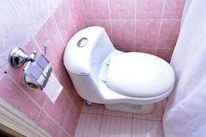 A white toilet with a bidet attachment and a paper towel dispenser in a bathroom with pink tiled walls. - Luxury One Hotel Lahore (Lahore)