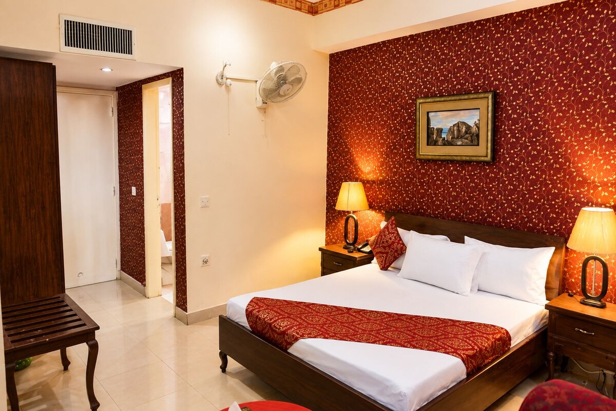Habitación de hotel con una cama grande, dos mesitas de noche con lámparas, una pared con diseño rojo, un cuadro enmarcado y un banco de madera.