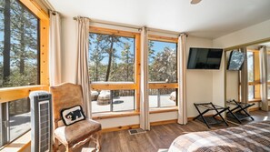 A cozy room with a bed, a chair with a pillow, a TV, and large windows offering a view of trees and a snowy landscape. - Dancing Bear Lodge, Pine AZ; Luxury Cabin with Views, Spa & Outdoor living area (Pine)