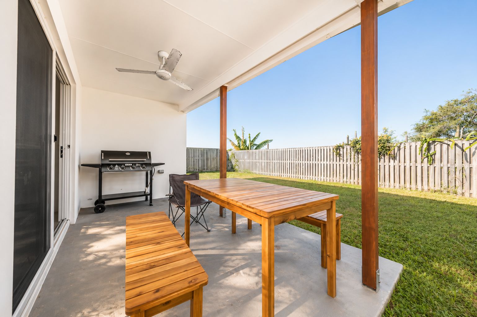 An outdoor patio with a wooden table and benches, a grill, and a ceiling fan.