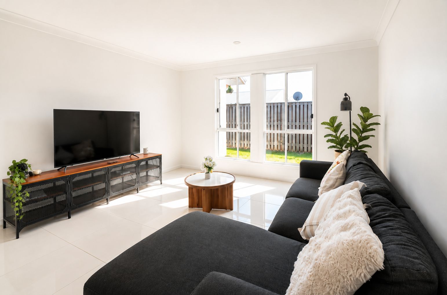 A modern living room with a flat-screen TV, a dark sofa, a round coffee table, and a large window with a view of a fence and greenery.