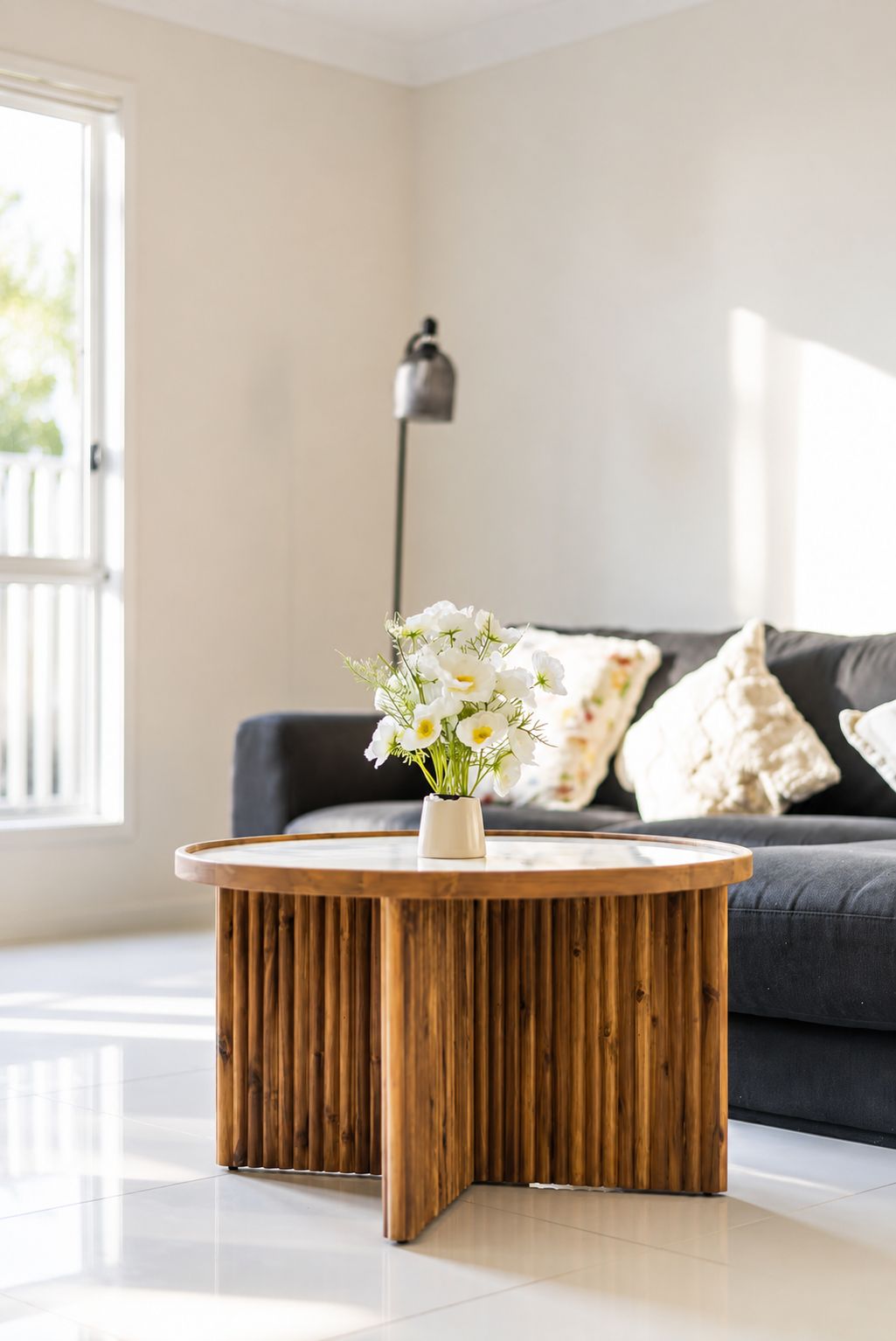 A round wooden coffee table with a vase of white flowers in a modern living room with a dark sofa and a lamp in the background.