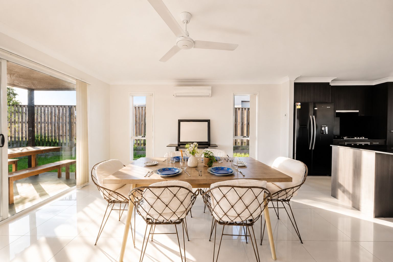 A modern kitchen with a built-in island, a dining area with a wooden table and wireframe chairs, and a sliding glass door leading to an outdoor patio.