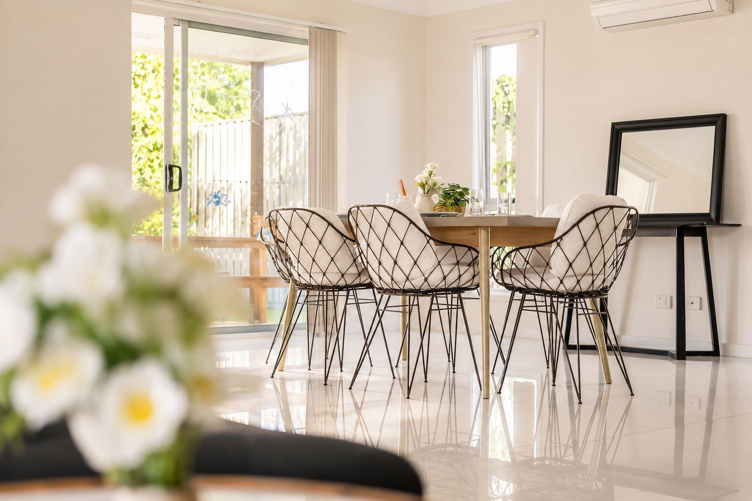 A modern dining area with a round table, four wireframe chairs, and a large mirror on the wall.