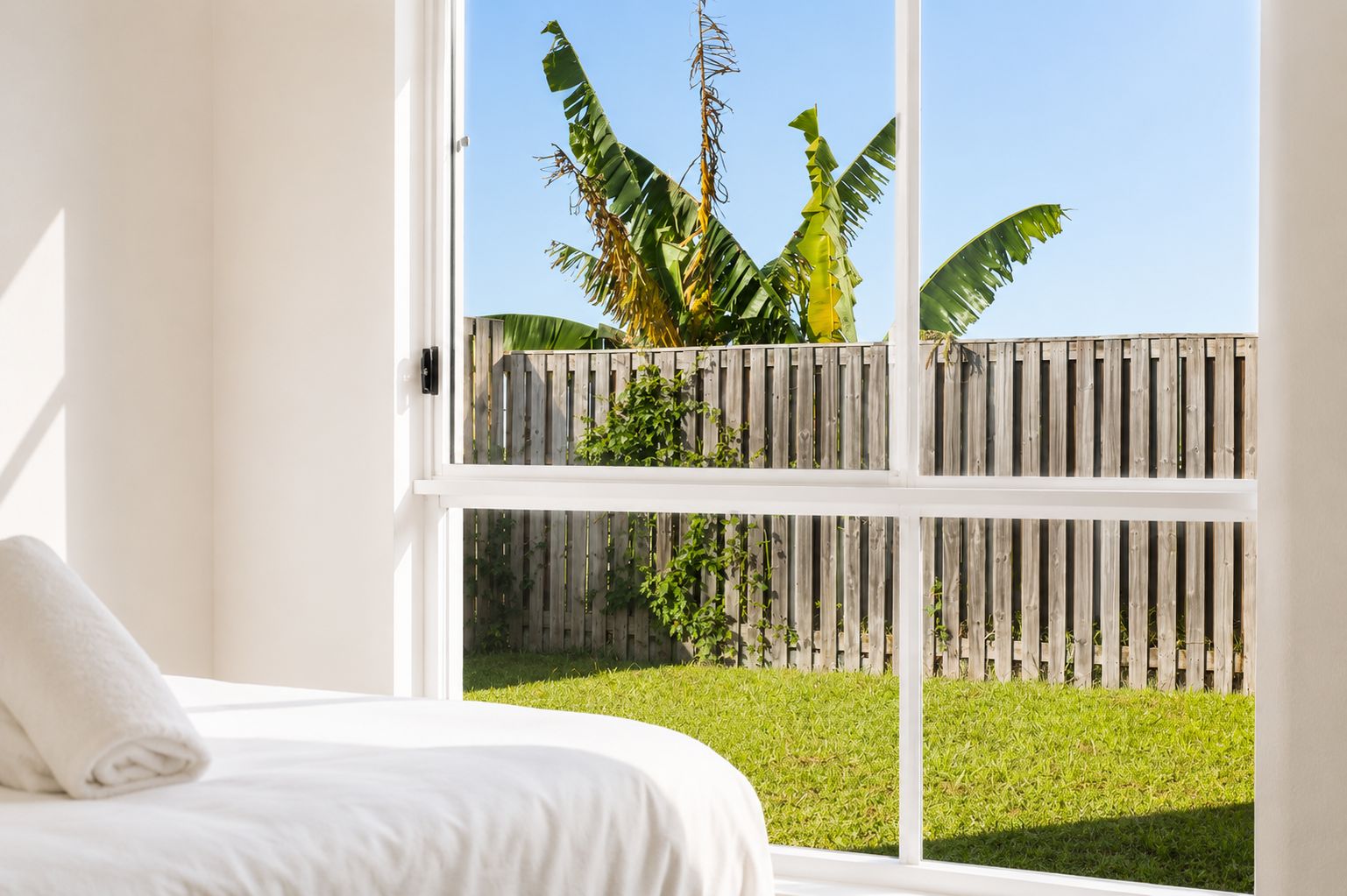 A bedroom with a large window overlooking a grassy yard and a wooden fence.