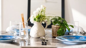 Taula de menjador preparada amb plats blaus, una gerra blanca amb flors, una planta verda en cistella i gots de vidre transparent.