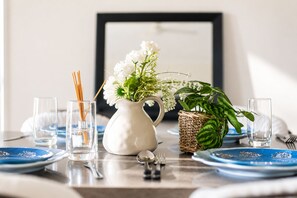 Ein Esstisch ist mit blauen Tellern, einer weißen Vase mit Blumen, einer grünen Pflanze in einem Korb und klaren Gläsern gedeckt.