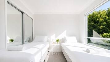 A modern bedroom with two beds, a small table with a plant, and a large window with a view of greenery.
