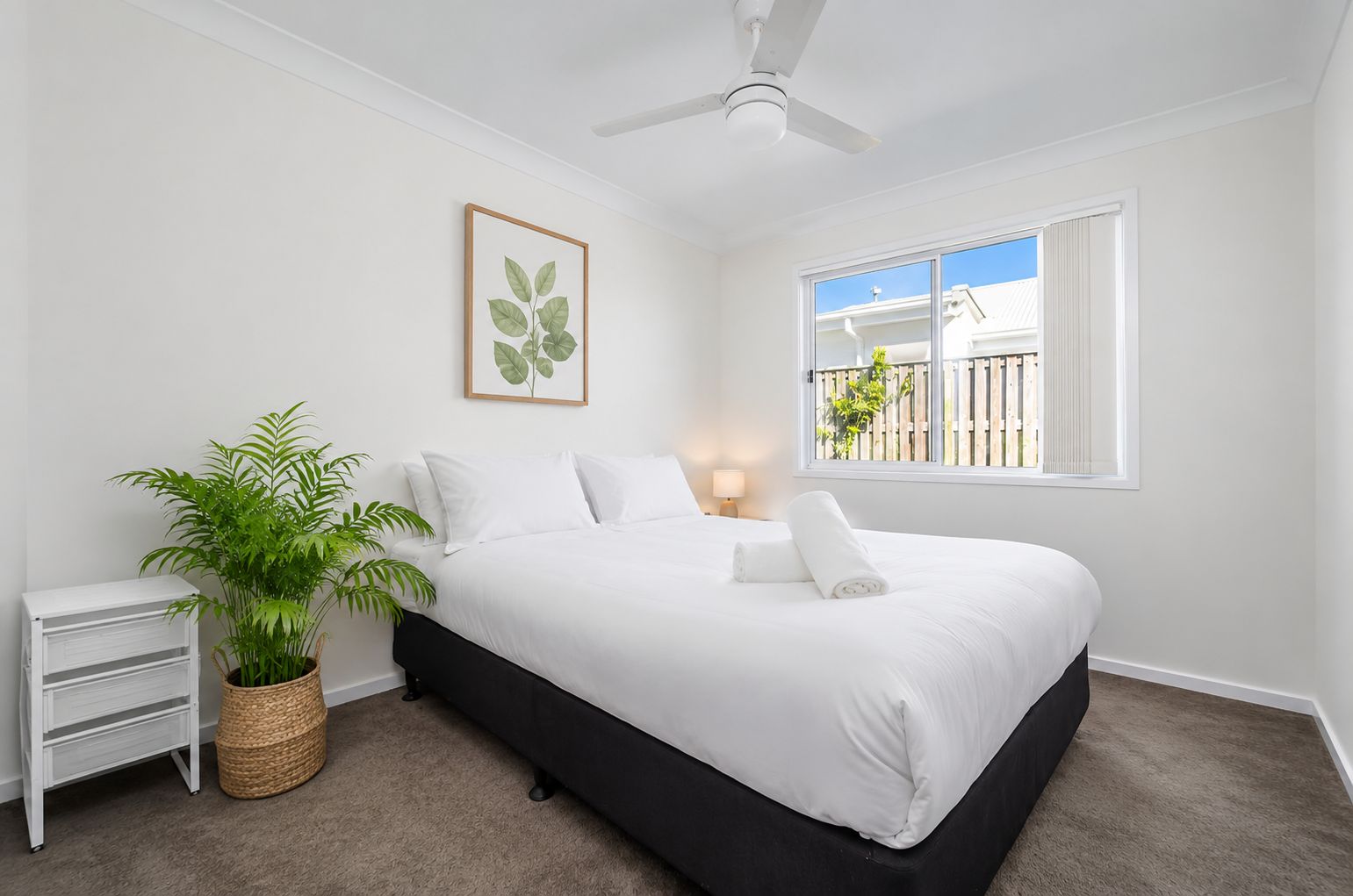A bedroom with a bed, a ceiling fan, a window with a view, a bedside table, and a potted plant.