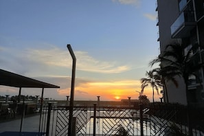 Auringonlaskunäkymä parvekkeelta, jossa on kaide, palmuja ja taustalla näkyvä rakennus.