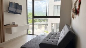 Ein modernes Wohnzimmer mit einem grauen Sofa, einem Wandfernseher und einem großen Fenster mit Blick auf ein Gebäude und Grünflächen.