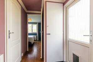 A narrow hallway with a bed in the background, a radiator, and a window with curtains.