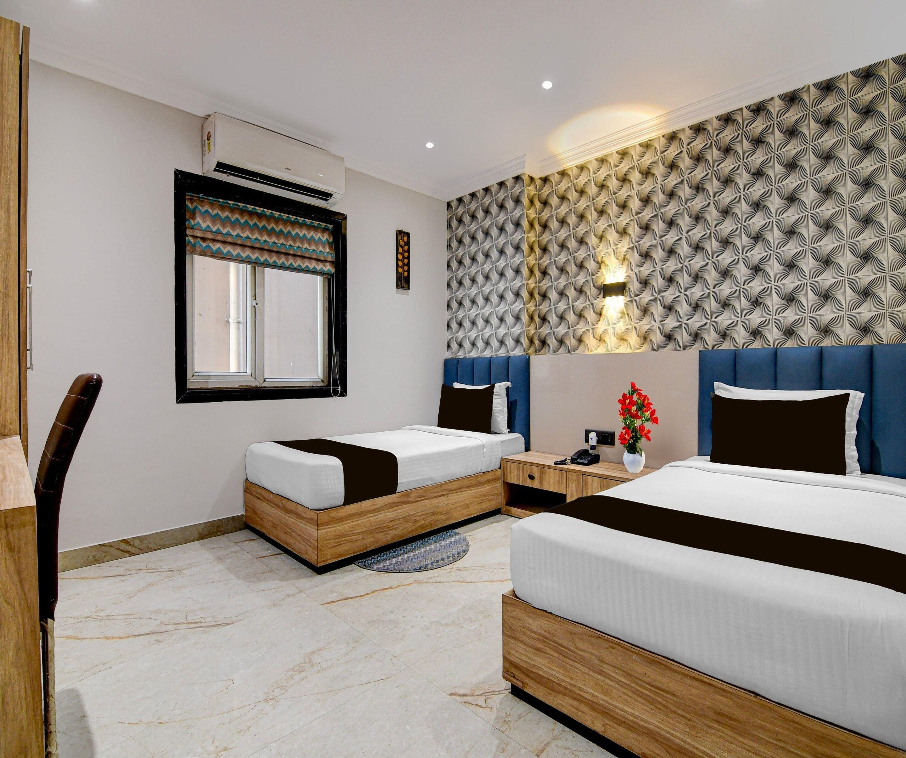 A hotel room with two beds, a desk, a chair, a window with a patterned curtain, and a wall with a geometric design.