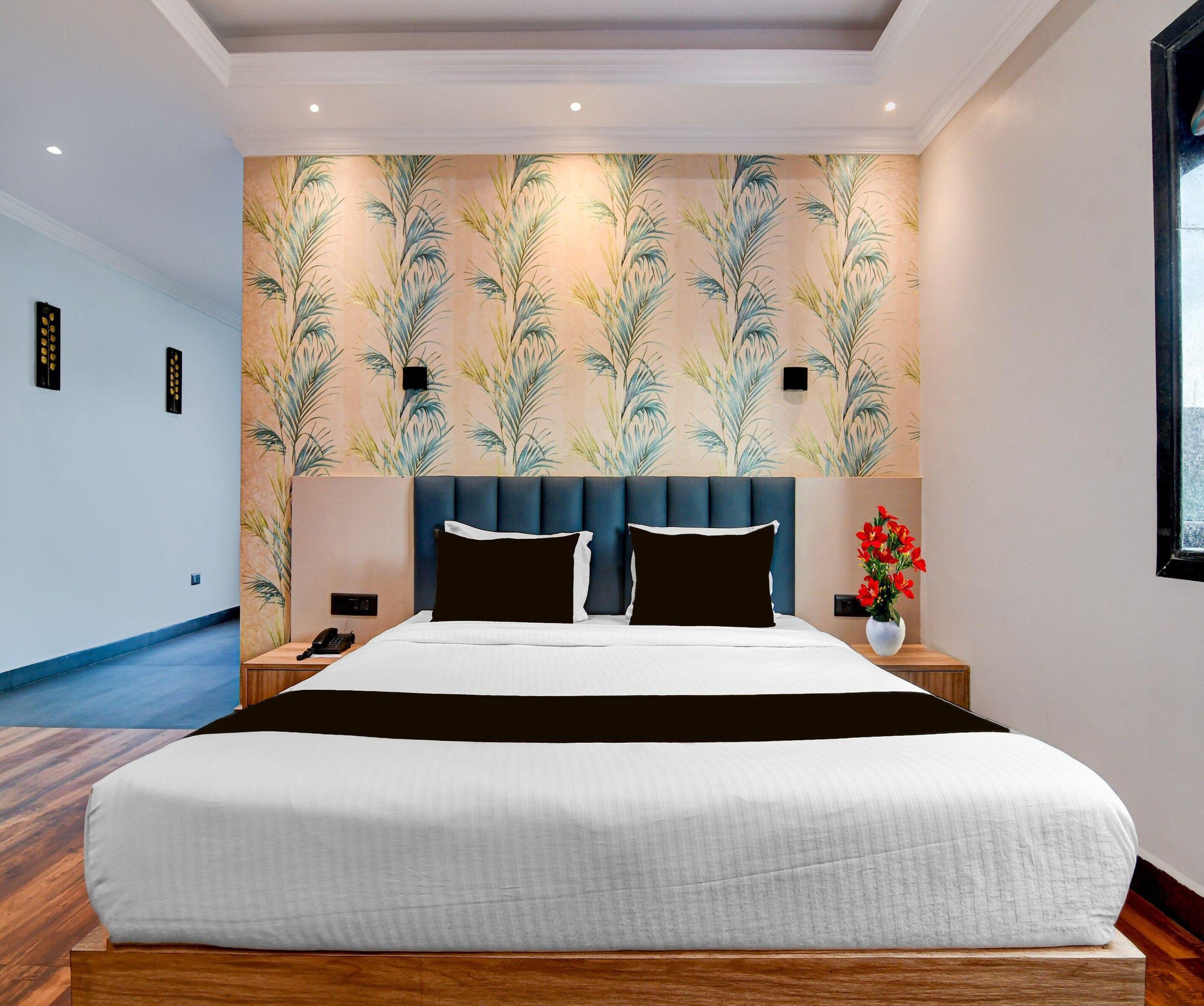 A hotel room with a large bed, a headboard, a nightstand with a phone, a wall with a palm leaf pattern, and a window.