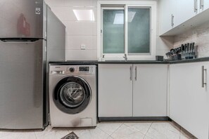 A compact kitchen with a stainless steel refrigerator, a front-loading washing machine, white cabinets, and a window above the sink. - HAVN - 1 Bedroom Apartment in G24 Tower, JVC (Dubai)