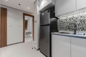 A modern kitchen with a stainless steel refrigerator, a sink with a mosaic backsplash, and a wooden door leading to another room. - HAVN - NEW 1 Bedroom in Neva Residences, JVC (Dubai)