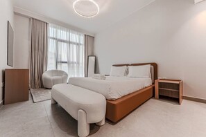 A modern bedroom with a large bed, a bedside table, a chair, and a small table. - HAVN - NEW 1 Bedroom in Neva Residences, JVC (Dubai)