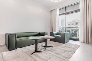 A modern living room with a green sofa, a round table, and a view of a cityscape through large windows. - HAVN - NEW 1 Bedroom in Neva Residences, JVC (Dubai)