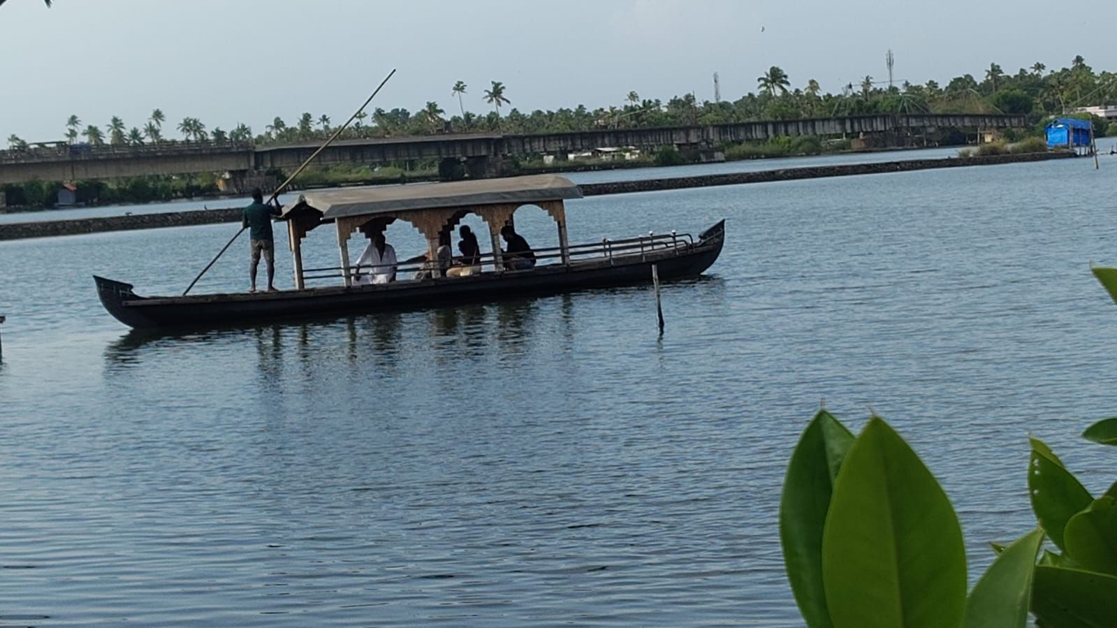 A traditional boat with a roof, people on board, and a calm water body with a bridge and greenery in the background.