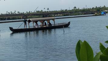 A traditional boat with a roof, people on board, and a calm water body with a bridge and greenery in the background.