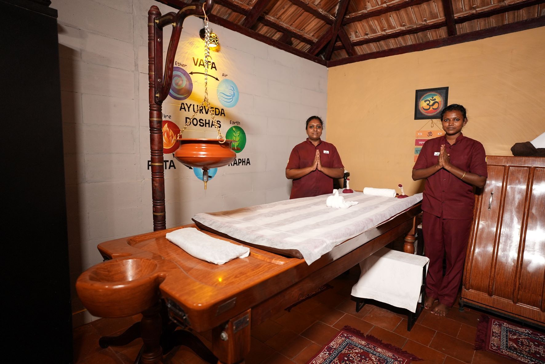 Een traditionele Ayurvedische therapieruimte met een houten tafel, een messing pot en twee therapeuten.