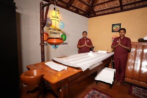 Een traditionele Ayurvedische therapieruimte met een houten tafel, een messing pot en twee therapeuten.