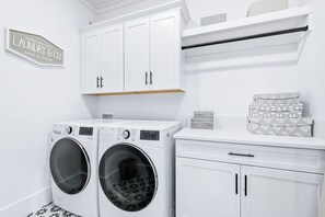 A laundry room with two front-loading washing machines, a white cabinet with black handles, and a sign indicating 24-hour service. - Luxury Home, Gourmet Kitchen, Sleeps 14, Balconies,Community Pool, Grayton Beach (Santa Rosa Beach)