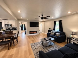 Ein modernes Wohnzimmer mit Kamin, einem Essbereich mit Holztisch und Stühlen, einem schwarzen Sofa und einem Deckenventilator.