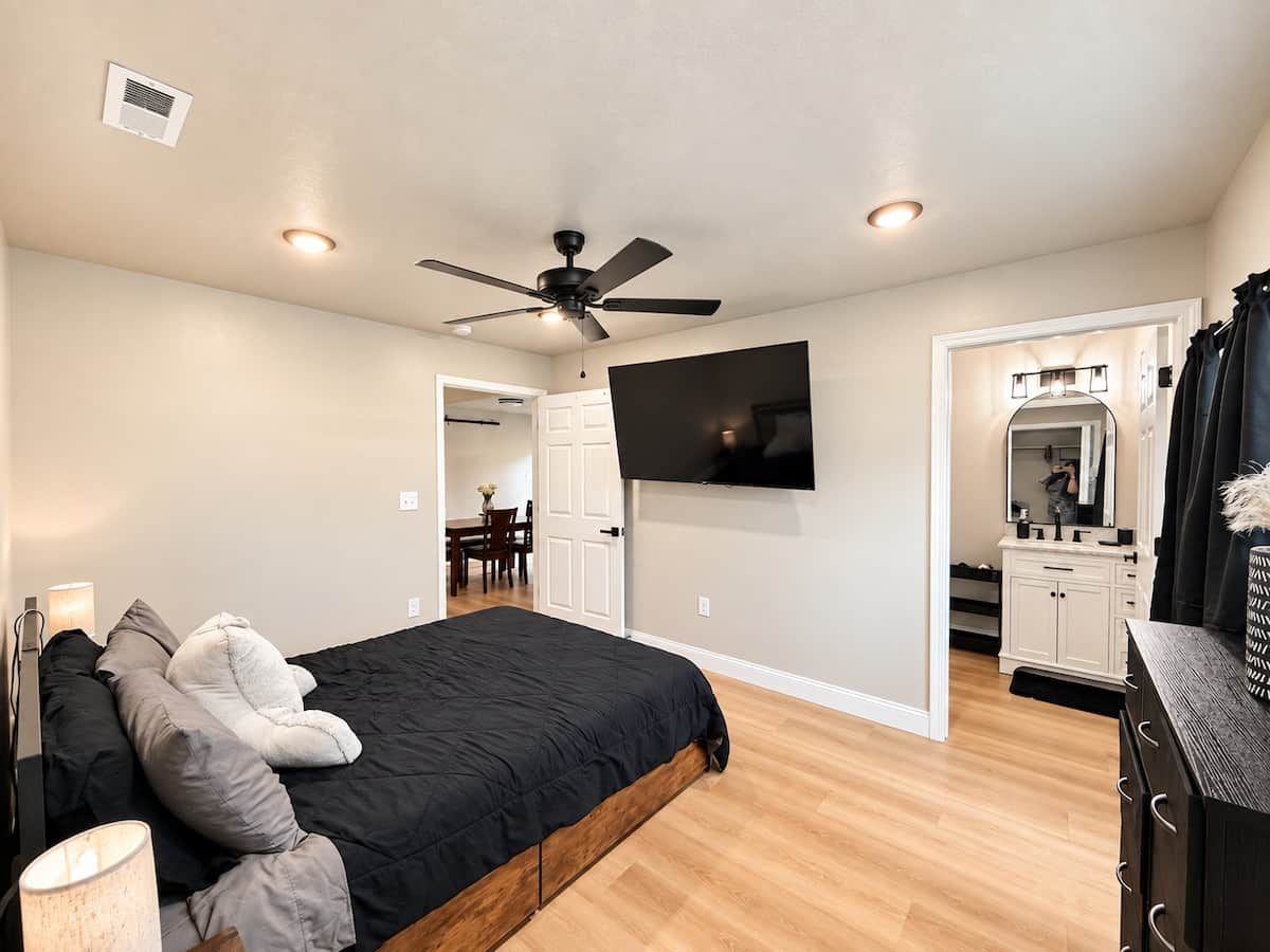 Une chambre à coucher avec un lit, un ventilateur de plafond, une télévision, une commode et une salle de bain visible à travers une porte ouverte.