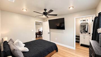 Une chambre à coucher avec un lit, un ventilateur de plafond, une télévision, une commode et une salle de bain visible à travers une porte ouverte.