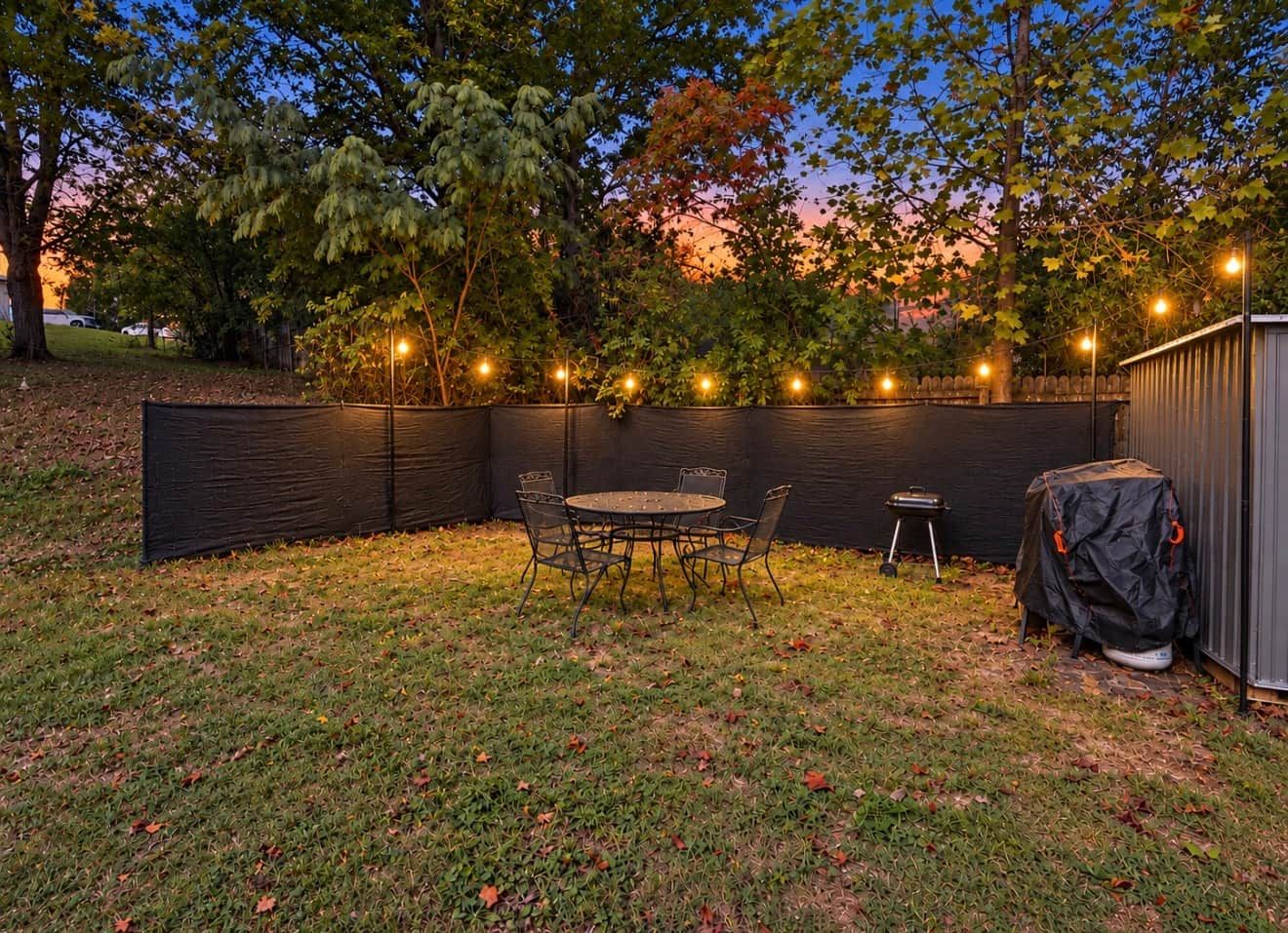 Een achtertuin met een overdekte patio tafel en stoelen, omringd door een zwarte hek en lichtslingers.