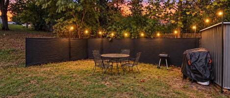 A backyard with a covered patio table and chairs, surrounded by a black fence, and string lights.