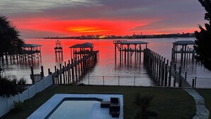 Piscina com vista para um pôr do sol sobre a água, cercada por docas de madeira e uma área gramada.