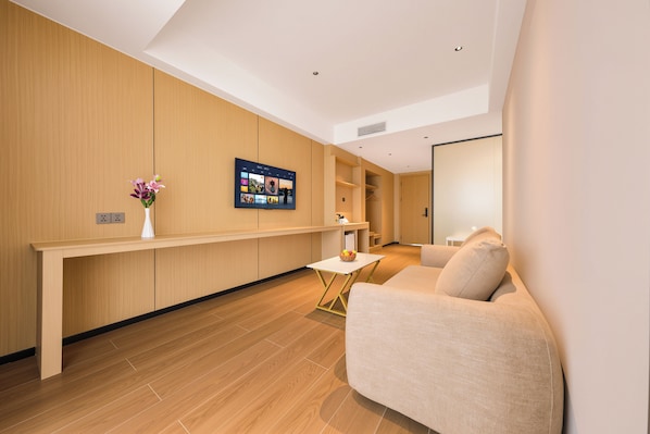 Una habitación de hotel moderna con suelo de madera, un sofá, una pequeña mesa con un cuenco de fruta y un televisor de pared que muestra una galería de fotos.
