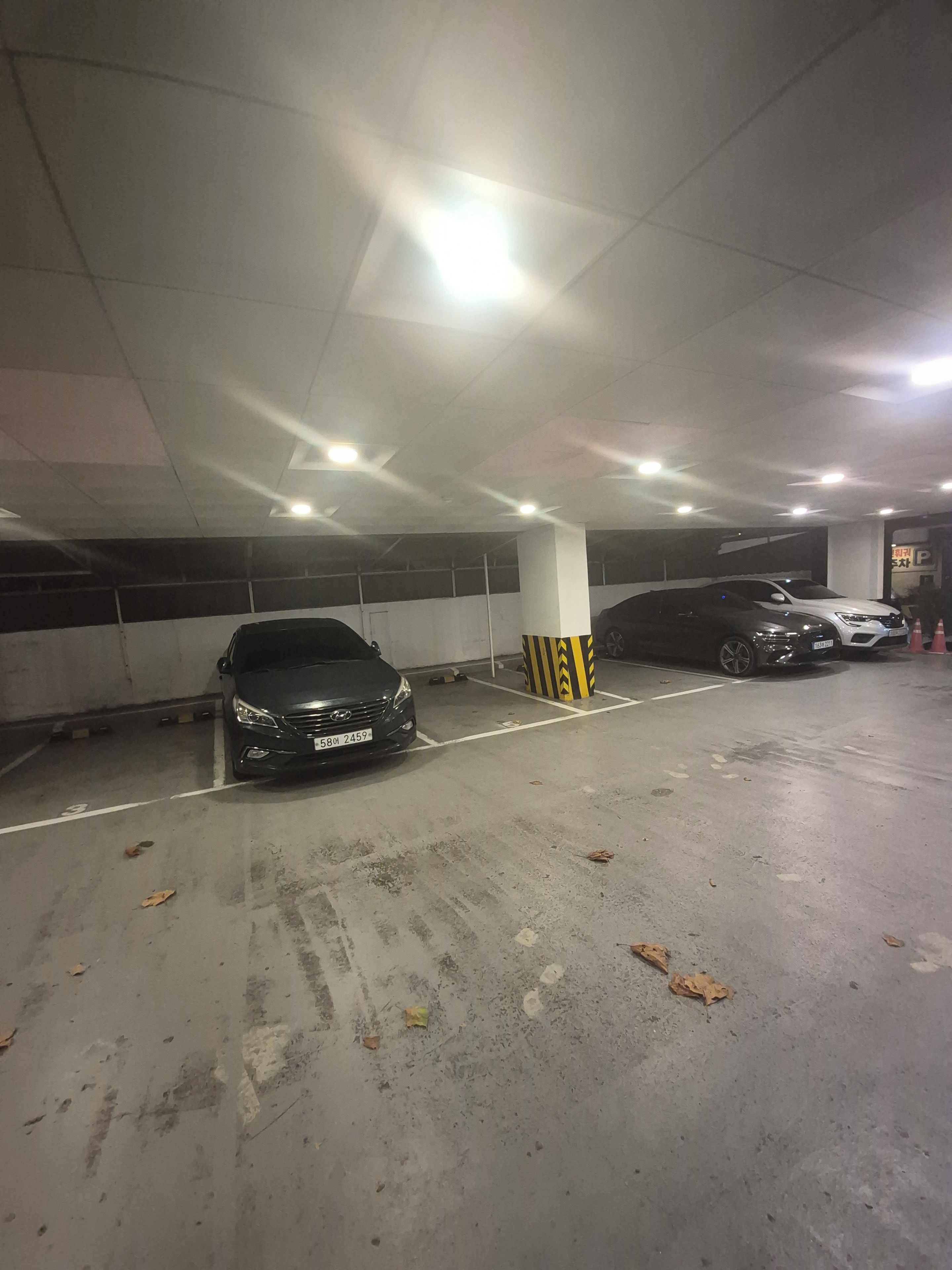 A parking garage with multiple cars parked, concrete pillars, and overhead lighting.