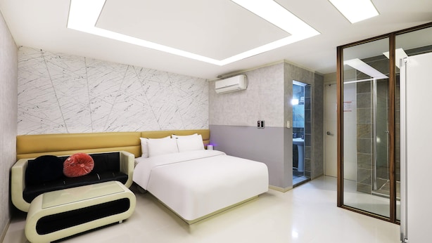 Quarto moderno com cama, sofá, banheiro com chuveiro de vidro e teto com iluminação embutida.