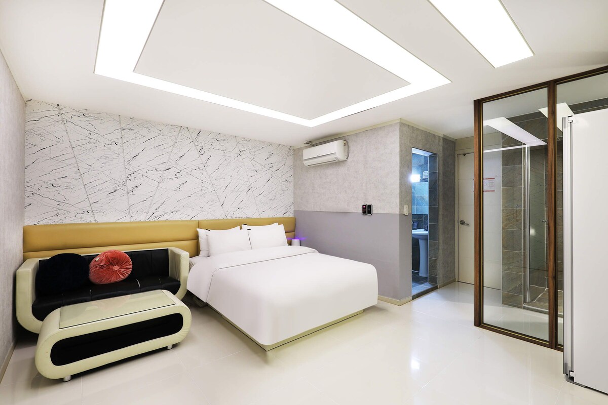Ein modernes Schlafzimmer mit Bett, Sofa, Badezimmer mit Glasdusche und einer Decke mit Einbauleuchten.