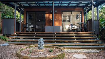 A modern building with a covered outdoor seating area, steps leading to a door, and a stone sculpture in the foreground.