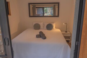 A neatly made bed with pillows and a folded towel, a bedside table with a lamp, and a window with a view of greenery.